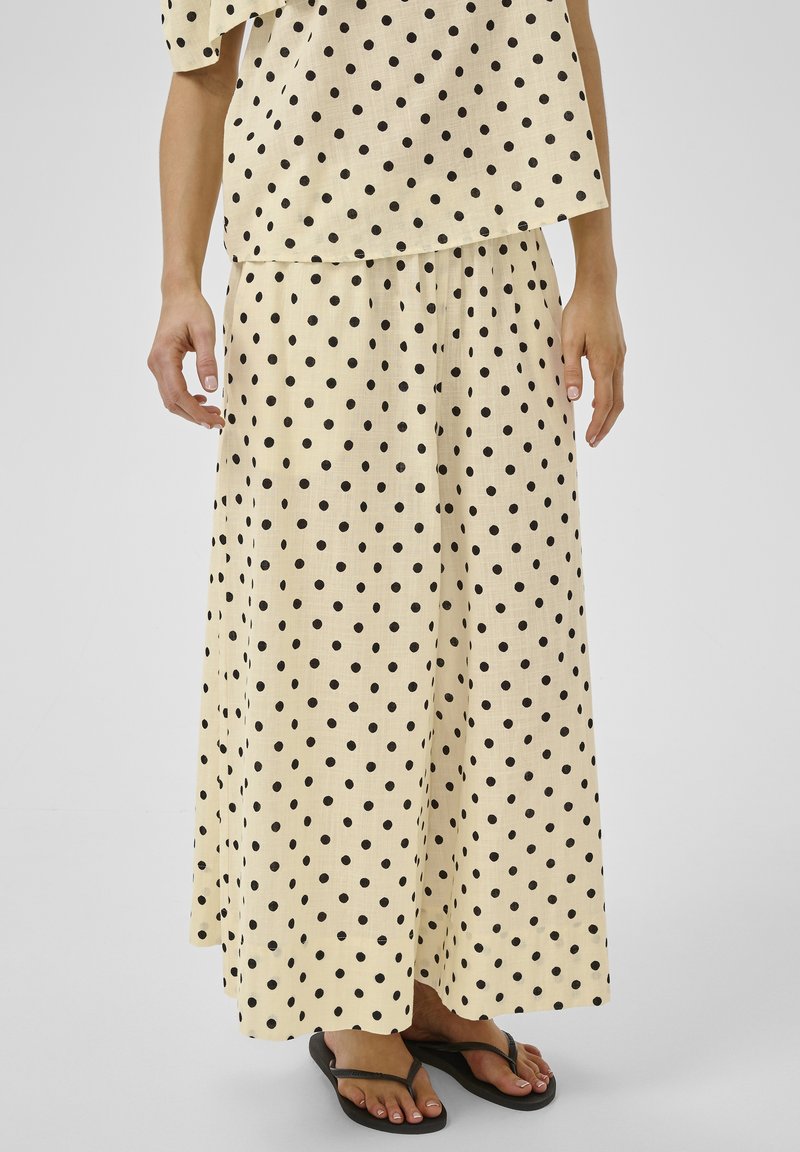Ensemble jupe beige et top avec des pois noirs, porté avec des tongs noires, montré de la moitié du torse jusqu'aux pieds sur un fond uni.