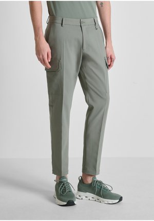 Uomo che indossa pantaloni cargo verde chiaro e sneakers verdi abbinate, in piedi contro uno sfondo semplice grigio chiaro.