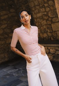 Noor naine lühikeste mustade juustega, kes kannab punase-valgete triipudega polo särki ja valgeid pükse, seisab kiviseina taustal.