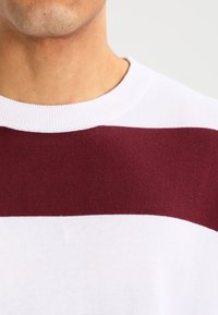 Pull blanc avec de larges rayures horizontales bordeaux, présentant un col rond côtelé et une texture en maille lisse.