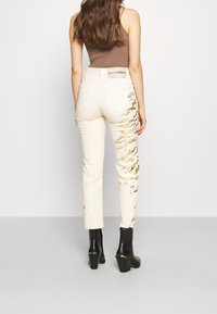 Jeans crema con detalles de salpicaduras de pintura dorada en la pierna izquierda, con una cintura de altura media y bolsillos traseros. Llevados con botines negros de tacón.