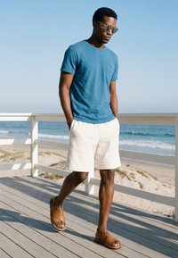 Man wearing blue t-shirt, white shorts, and brown sandals walking on a wooden deck by the beach with ocean waves in the background.