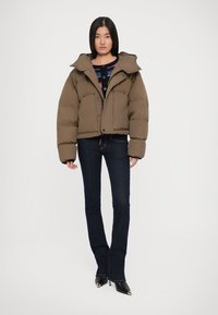 Doudoune marron avec un col montant, fermeture éclair et pression, et quatre poches avant. Portée par-dessus un pull à motifs et associée à un jean foncé.