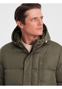 Olivengrøn pufferjakke med hætte, frontlynlås og stitched paneler. Har en brystlomme og en mat finish tekstur.