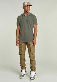 Polo di colore verde oliva con maniche corte, abbinato a pantaloni cargo beige con molte tasche. Sneakers alte bianche e grigie. Cappellino color beige.