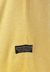 Camiseta de tela amarilla con un acabado texturizado. Presenta una etiqueta negra con texto blanco que detalla la marca y el eslogan a lo largo de la costura lateral.