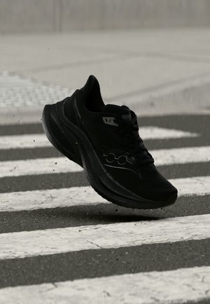 Chaussure de sport noire capturée en plein air au-dessus d’un passage piéton sur asphalte avec de petits débris soulevés autour.