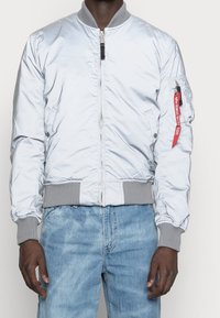 Light blue bomber jacket with a zip front, ribbed collar and cuffs, and a pocket on the arm featuring a red pull tab. Textured finish.