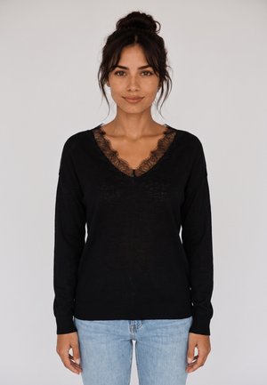 Donna con capelli scuri raccolti in uno chignon, che indossa un maglione nero a scollo a V con rifiniture in pizzo e jeans azzurri, in piedi davanti a uno sfondo chiaro uniforme.