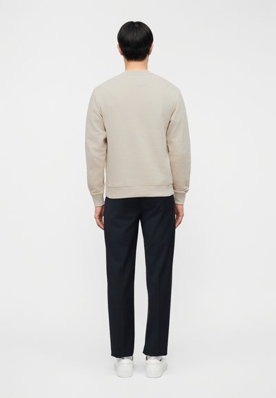 Μπεζ φούτερ με στρογγυλή λαιμόκοψη και ριμπ μανσέτες, σε συνδυασμό με σκούρο μπλε παντελόνι και λευκά αθλητικά παπούτσια, εμφανίζεται από πίσω με φόντο απλή επιφάνεια.