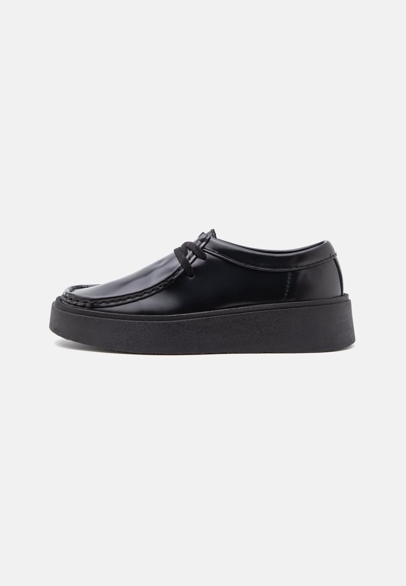 Chaussure en cuir noir brillant avec une semelle épaisse en caoutchouc noir et des lacets noirs, vue de côté sur un fond clair uni.