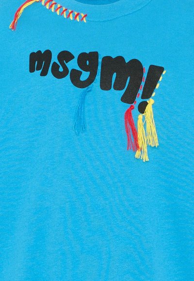 Tela azul con texto negro "msgm!", decorada con borlas cosidas en rojo, amarillo y azul cerca del cuello y del signo de exclamación.