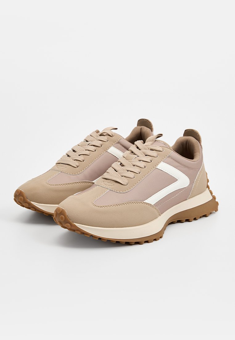 Zapatillas de deporte beige y rosa con un diseño texturizado, que presentan acentos blancos, paneles de malla y una suela de goma con forma redondeada.