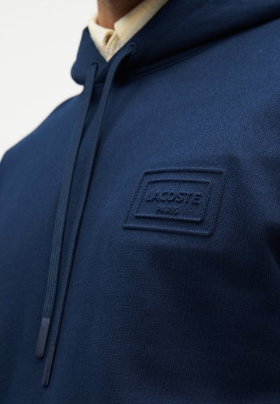 Homme portant un sweat à capuche Lacoste bleu marine avec cordons de serrage et un patch logo en relief sur la poitrine, par-dessus une chemise beige à col.