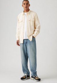 Lichtbeige knoopshirt met twee borstzakken, gedragen over een wit T-shirt, gecombineerd met lichtblauwe jeans en zwarte sneakers met camouflageaccenten.