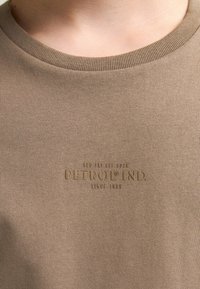 T-shirt in cotone marrone a coste con collo rotondo. Presenta il logo in rilievo "PETROL IND. dal 1989" al centro. Texture liscia.
