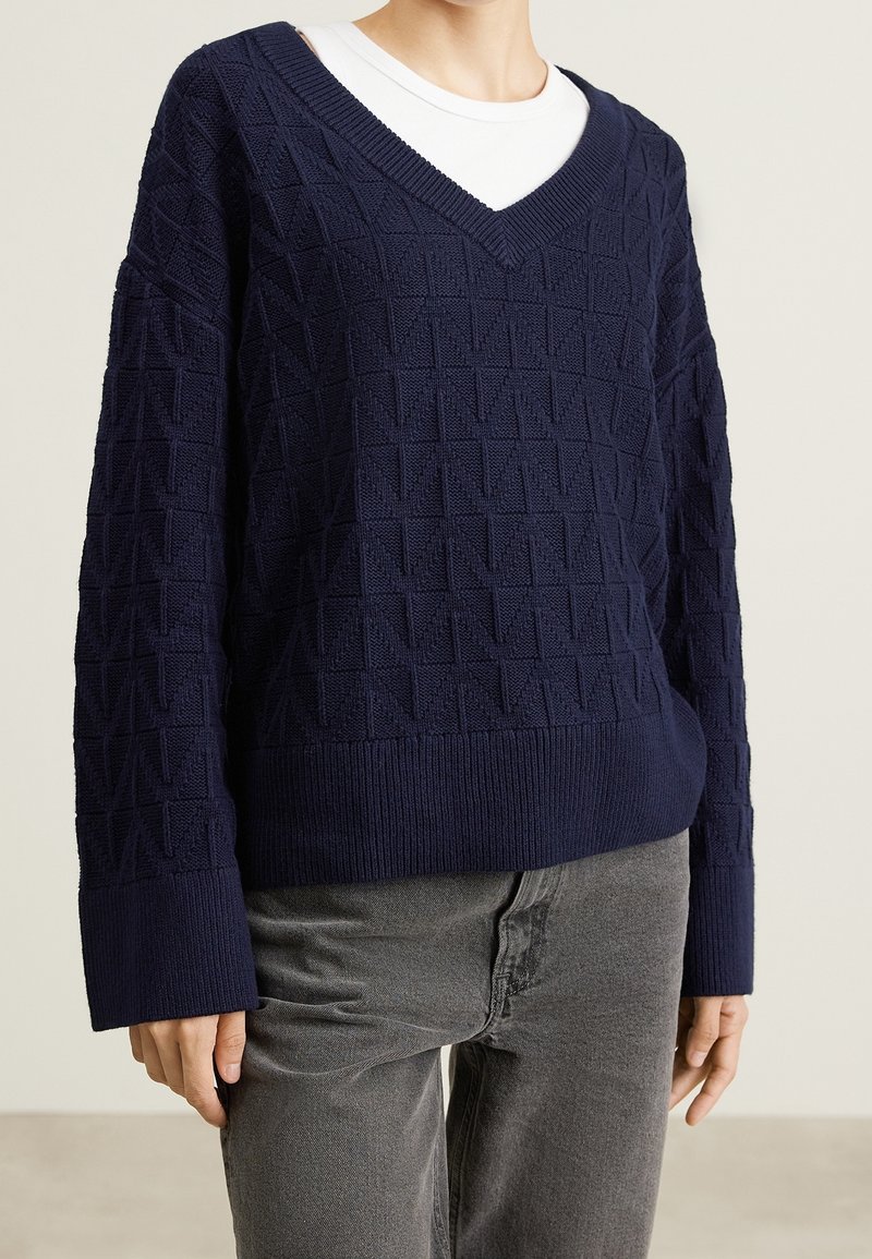 Femme portant un pull à col en V texturé bleu marine par-dessus un T-shirt blanc et un jean gris, debout devant un fond clair uni.