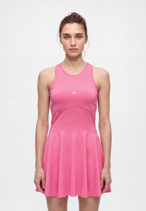 Une femme se tient face à l’objectif, vêtue d’une robe de tennis rose vif sans manches, sur un fond blanc uni de studio.