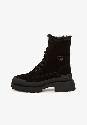 Osoba nosząca czarne masywne buty na platformie z futrzaną wyściółką, białe grube dzianinowe ochraniacze na nogi oraz czarne legginsy, stojąca na białej podłodze.