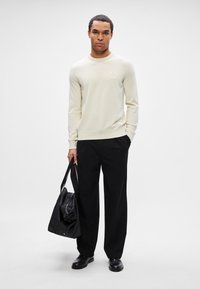 Mann in cremefarbenem Langarmpullover und schwarzen Hosen, der eine schwarze Tragetasche hält, vor einem weißen Hintergrund stehend.