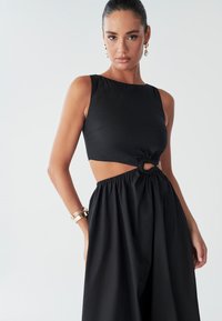 Model draagt een stijlvolle zwarte outfit met een uitsnijding en een geplooide rok. Ze poseert zelfverzekerd tegen een effen achtergrond.