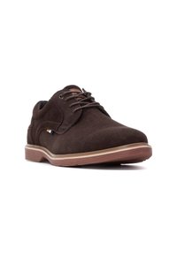 Zapatos de ante marrón con puntera redondeada, diseño de cordones y suela de goma. Presenta un pequeño acento en azul y amarillo en el lateral.
