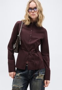 Νέα γυναίκα που φορά γυαλιά με σκιασμένους ορθογώνιους φακούς, σκούρο μαροτίνι πουκάμισο με κουμπιά και ζώνη στη μέση, σκισμένο τζιν με μεταλλικά τρουκ, και τσάντα στον ώμο.