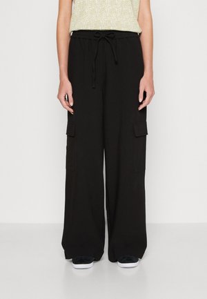 Osoba ubrana w czarne, szerokie spodnie bojówki z ściągaczem w pasie i klapkowymi kieszeniami, w połączeniu z czarno-białymi butami i jasną bluzką.