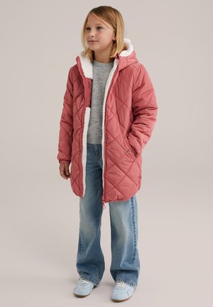 Niña joven de pie con un abrigo acolchado rosa con capucha y forro de felpa blanca, suéter gris, jeans azules y zapatillas azul claro sobre un fondo beige.