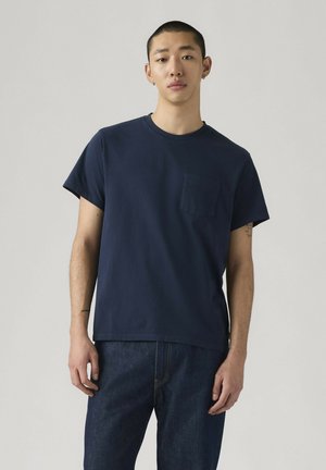 Jonge man met kort haar, gekleed in een navyblauw T-shirt met korte mouwen en een borstzakje, en donkere blauwe spijkerbroek, staand tegen een effen achtergrond.