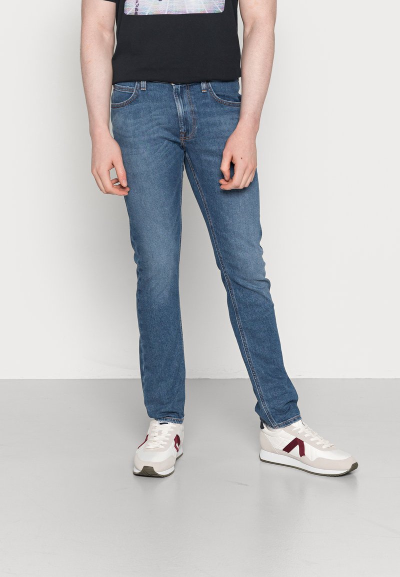 Jeans de denim azul de corte slim com um leve desgaste, combinados com ténis brancos com detalhes em bordô e uma sola texturizada.