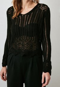 Schwarzer Strickpullover mit transparentem Häkeldesign, langen Ärmeln und gewelltem Saum, der ein strukturiertes Muster im gesamten Pullover hervorhebt.