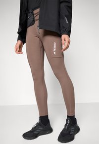 Brune, tettsittende leggings laget av elastisk stoff, med sømdetaljer og en sidelomme med en hvit logo. Kombinert med svarte treningssko.