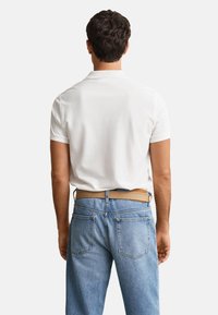 Man with short dark hair wearing a white short-sleeve polo shirt and light blue jeans with a tan belt, facing away from the viewer.