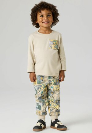 Smiling child with curly hair wears beige long-sleeve shirt and pants with colorful dinosaur comic strip prints, standing against plain background.