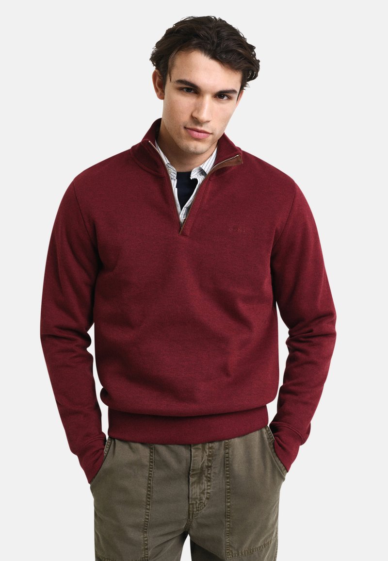 Maglione pullover bordeaux con mezza zip, con finitura testurizzata e polsini a coste, abbinato a pantaloni khaki. Presenta un logo semplice sul petto.