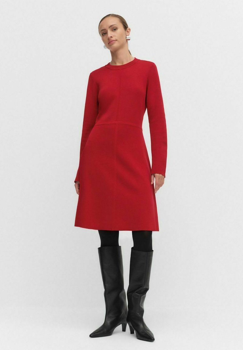 Rode jurk met lange mouwen, een aansluitend lijfje en een wijduitlopende rok, gemaakt van zachte stof. Gecombineerd met zwarte kniehoge laarzen en donkere panty’s.