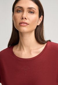 Rode kortemouwtop met een ronde halslijn en een decoratief strassdesign dat in het midden vooraan op een boom lijkt. Glad textuur.