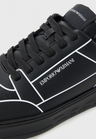 Baskets noires avec accents géométriques blancs, bout rond, surface texturée et logo sur le côté. Comprend un système de laçage noir et une semelle plate.
