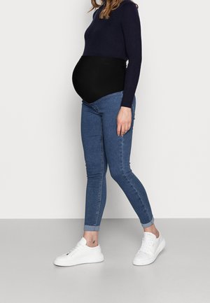 Zwangere persoon draagt een zwarte zwangerschapsband, een marineblauwe trui, blauwe spijkerbroek en witte sneakers, staand tegen een effen achtergrond.