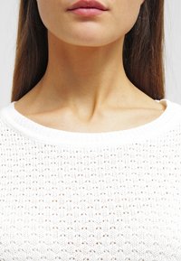 Vit stickad tröja med rund halsringning, texturerat mönster med små öppna vävningar och ribbat avslut vid kragen och ärmarna.