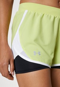 Närbild av en person som bär ljusgröna sportshorts med vit kant och svarta innerbyxor, med handen vilande bredvid låret.