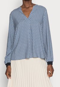 Frau in einer blauen und weißen geometrisch gemusterten Bluse mit langen Ärmeln und schwarzen Bündchen, dazu einen weißen Faltenrock.