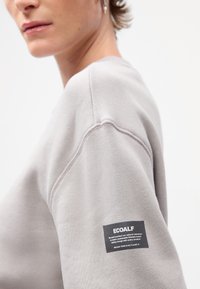 Lys grå sweatshirt med rund hals, ribbede ermer og et svart Ecoalf-merke på armen med hvit tekst. Myk tekstur.