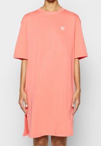 Volný oversized tričkový šaty světle korálové barvy z bavlny, s kulatým výstřihem, krátkými rukávy a malým bílým logem Adidas na hrudi.