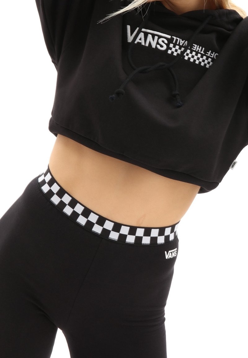 Femme portant un sweat à capuche court noir Vans et des leggings noirs avec une taille à carreaux noir et blanc et le logo Vans.