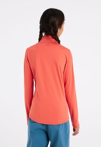Langermige koraalkleurige top van soepel, rekbaar materiaal met een hoge kraag en vlakke naden; gecombineerd met blauwe broek en van achteren gedragen.