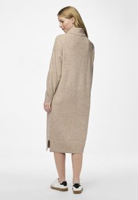 Robe longue beige en matériau doux avec un col haut, des poignets côtelés, un détail d'ouverture sur le côté et une coupe ample. Associée à des baskets blanches.