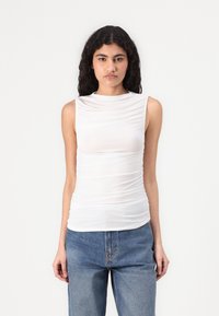 White sleeveless top with gathered texture, high neckline, and a slim fit, paired with loose blue jeans. Smooth fabric with slight sheen.