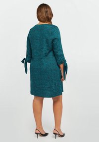 Tealfärgad kl änning med ett texturerat prickmönster, trekvartslånga ärmar och sidband. Designen är rak och faller ovanför knät.